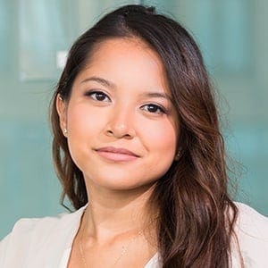 A woman with long brown hair, wearing a white top, is looking at the camera with a slight smile. The softly blurred background in cool tones hints at locations like Hunterdon or Mercer County NJ & Bucks County PA, home to trusted paving contractors.