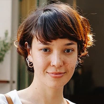 A person with short, wavy brown hair and light skin smiles softly at the camera. They are wearing hoop earrings and a white top, with a blurred background behind them in Mercer County NJ & Bucks County PA.