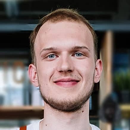 A young man with short light brown hair and a trimmed beard, smiling at the camera. Indoors, blurred shelves and lights in the background hint he's a paving contractor in Hunterdon, Mercer County NJ & Bucks County PA.