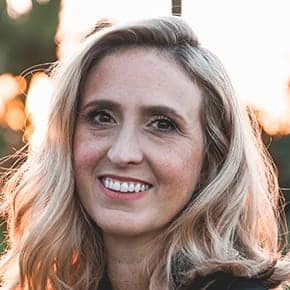 A woman with wavy blonde hair smiles at the camera outdoors, soft sunlight and trees blurred behind her—a welcoming face for a paving contractor serving Hunterdon, Mercer County NJ & Bucks County PA.