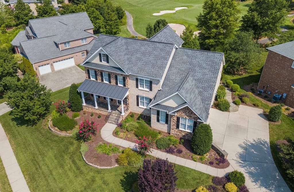 Aerial view of a large two-story brick house with a manicured yard, colorful shrubs, and curved driveway in a suburban neighborhood near a golf course, conveniently located in Mercer County NJ & Bucks County PA.