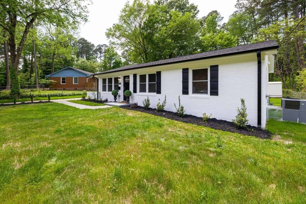 Single-story white brick house with black shutters, large front windows, neatly trimmed bushes, and a green lawn. A paved walkway by a trusted paving contractor in Hunterdon, Mercer County NJ & Bucks County PA leads to the front door. Trees and another house are visible behind.