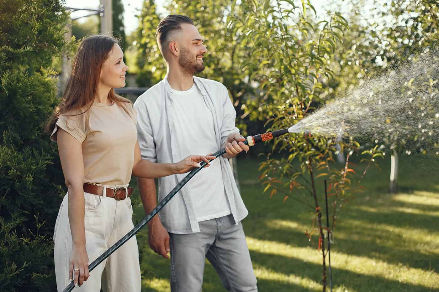 A woman and a man stand outdoors in a garden, watering plants with a hose together. They are smiling, surrounded by green trees and sunlight—enjoying their beautiful yard installed by a paving contractor in Hunterdon, Mercer County NJ & Bucks County PA.