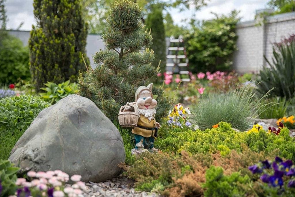A garden scene with a gnome statue holding a basket, standing next to a large rock and small tree, surrounded by colorful flowers and green shrubs—crafted by a paving contractor in Hunterdon, Mercer County NJ & Bucks County PA. A blurred fence completes the backdrop.