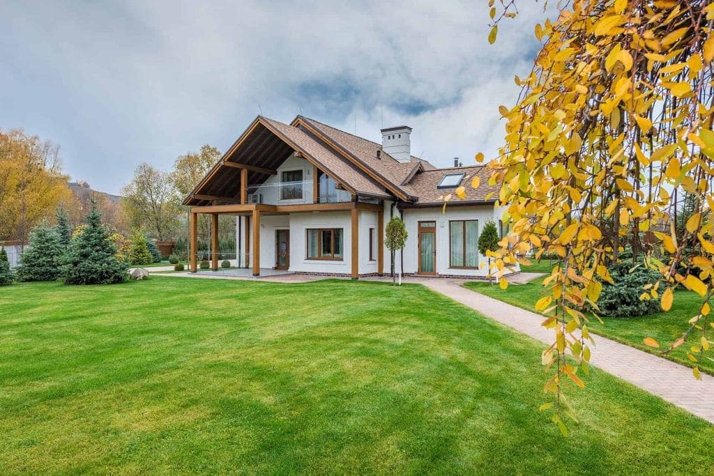 Modern two-story house with large windows and a wooden roof sits on a spacious, green lawn. Autumn trees with yellow leaves frame the foreground under a cloudy sky—perfect for residents of Mercer County NJ & Bucks County PA seeking serene living.