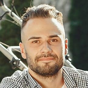 A man with short, neatly styled hair and a beard smiles at the camera. He is wearing a black and white checkered shirt and stands outdoors in Mercer County NJ, with greenery and a bicycle in the background.