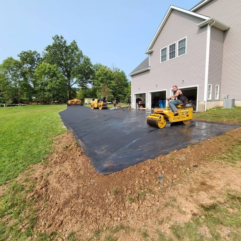 A paving contractor Hunterdon operates a yellow roller machine to smooth freshly paved asphalt on a driveway beside a gray house, with green grass and trees in the background in Mercer County NJ & Bucks County PA.