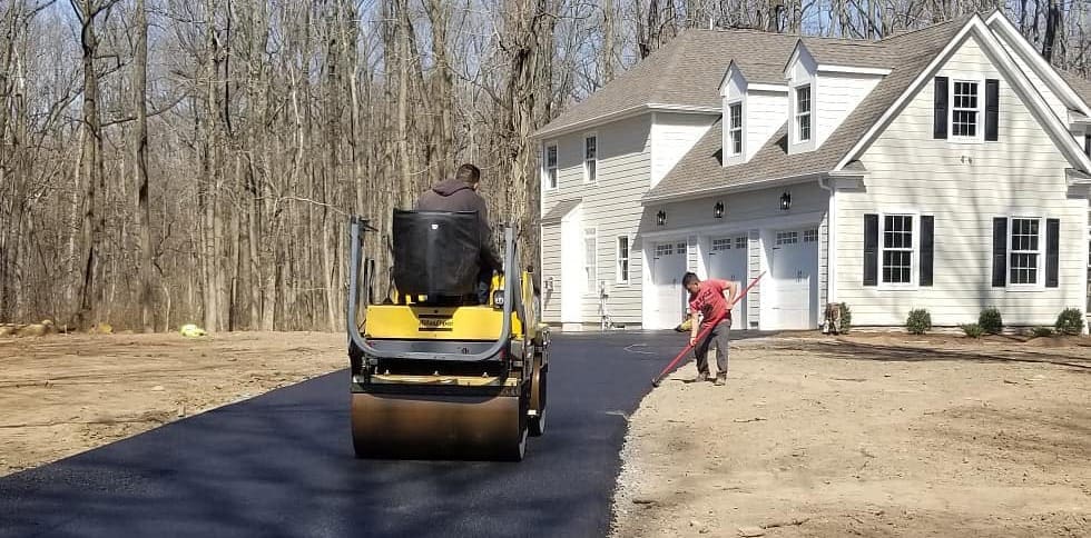 Two workers from a paving contractor Hunterdon pave a driveway in front of a white house; one operates a steamroller while the other levels the edge. Bare trees and dirt surround the freshly laid asphalt in Mercer County NJ & Bucks County PA.