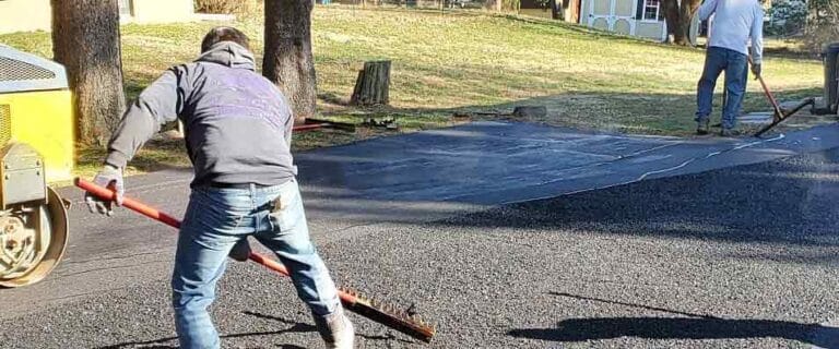 Two workers from a paving contractor in Hunterdon are paving a driveway; one spreads asphalt with a rake while the other smooths the surface. Trees and grass are visible in the background, typical of Mercer County NJ & Bucks County PA neighborhoods.
