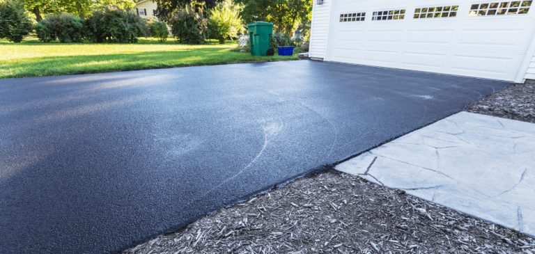 Freshly sealed black asphalt driveway in front of a white garage, bordered by mulch and concrete walkway, completed by a paving contractor Hunterdon, with a green lawn and trash bins in the background on a sunny day.