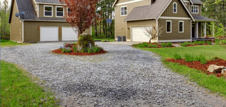 A gravel driveway curves between a two-story house and detached garage, both brown with white trim. Green grass, landscaped plants, and trees surround the property—perfect for upgrades by a paving contractor in Hunterdon, Mercer County NJ & Bucks County PA.