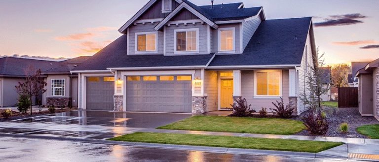 A modern two-story house with gray siding, large windows, and a double garage. In Mercer County NJ & Bucks County PA, the front yard's green lawn and wet pavement—possibly done by a paving contractor in Hunterdon—reflect sunset colors after rain.
