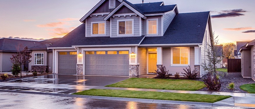 A modern two-story house with gray siding, large windows, and a double garage. In Mercer County NJ & Bucks County PA, the front yard's green lawn and wet pavement—possibly done by a paving contractor in Hunterdon—reflect sunset colors after rain.