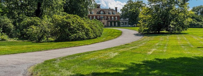 A curved driveway paved by a trusted paving contractor Hunterdon winds through a large, green lawn with trees on both sides, leading toward a spacious brick mansion under a partly cloudy sky.