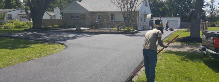 A worker smooths fresh asphalt on a driveway in front of a single-story house, while two other people from a paving contractor Hunterdon work in the background near a garage on a sunny day.