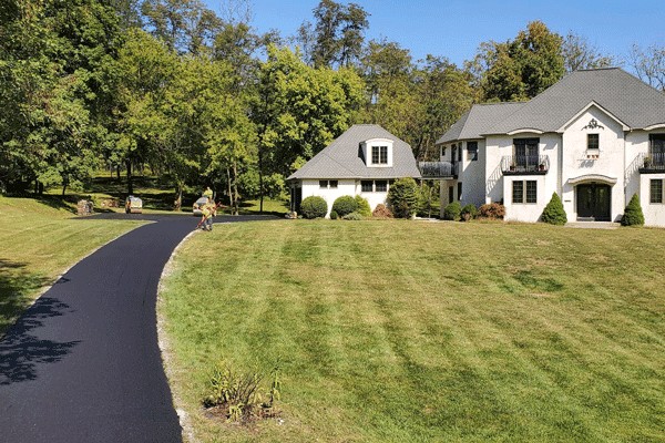 A paving contractor from Hunterdon operates a steamroller to pave a driveway leading to a large house, with construction equipment, gravel, and mounds of dirt visible on the grassy landscape in Mercer County NJ & Bucks County PA.
