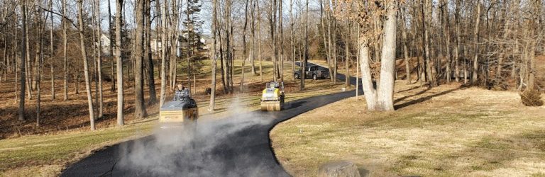 Two steamrollers pave a black asphalt driveway through leafless trees. Steam rises from the fresh pavement on a sunny day. A parked car sits by a house, showcasing the work of a paving contractor serving Hunterdon, Mercer County NJ & Bucks County PA.