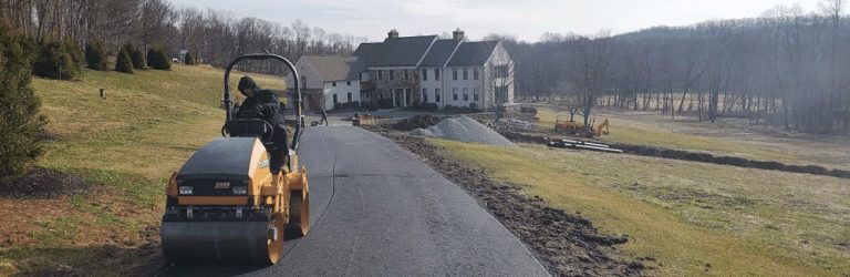 A paving contractor from Hunterdon operates a steamroller to pave a driveway leading to a large house, with construction equipment, gravel, and mounds of dirt visible on the grassy landscape in Mercer County NJ & Bucks County PA.