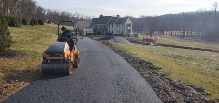 A paving contractor Hunterdon operates a steamroller, paving a black asphalt driveway to a large house surrounded by grass, trees, and construction equipment on a sunny day in Mercer County NJ & Bucks County PA.