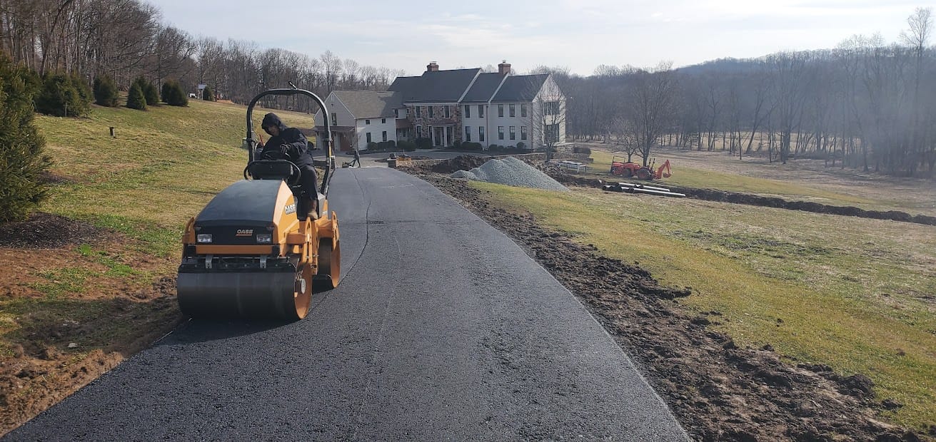 A paving contractor Hunterdon operates a steamroller, paving a black asphalt driveway to a large house surrounded by grass, trees, and construction equipment on a sunny day in Mercer County NJ & Bucks County PA.