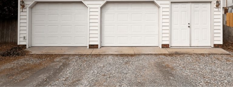 Two white garage doors and a single white entry door on a white building with gravel and a concrete driveway in front, expertly installed by a trusted paving contractor in Hunterdon, Mercer County NJ & Bucks County PA.