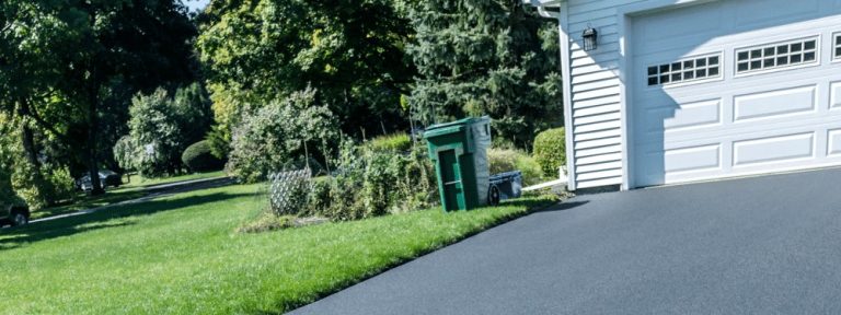 A freshly paved asphalt driveway by a top paving contractor in Hunterdon, Mercer County NJ & Bucks County PA leads to a white garage. Green grass, trees, and bushes surround the area, with a green trash bin and recycling bin near the garage.