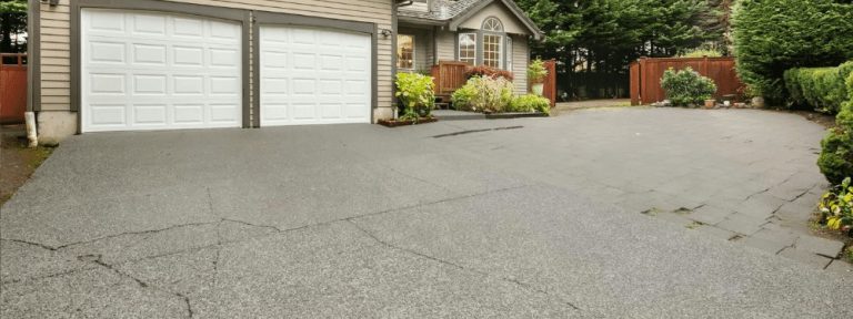 A wide concrete driveway with some cracks leads to a house with double white garage doors, lush green shrubs, potted plants, and a wooden fence—perfect for a paving contractor in Hunterdon, Mercer County NJ & Bucks County PA to restore.