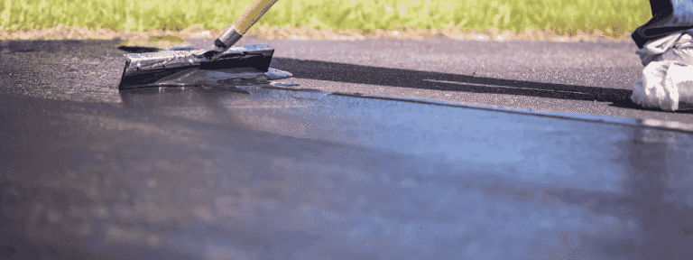 A person applies a fresh layer of black sealcoat to an asphalt driveway using a long-handled squeegee; green grass is visible in the background, showcasing the quality work of a paving contractor serving Mercer County NJ & Bucks County PA.