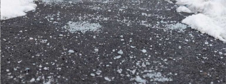 Close-up of an asphalt road in Mercer County NJ & Bucks County PA, scattered with ice melt or rock salt and patches of snow. The wet, cold surface highlights winter weather—perfect for a paving contractor Hunterdon to ensure safe roads.