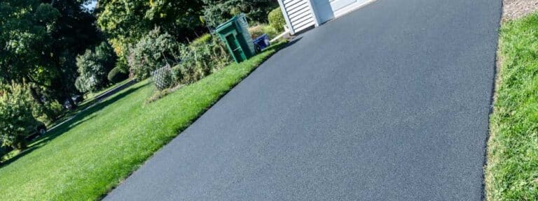 Freshly paved black asphalt driveway by a paving contractor in Hunterdon, Mercer County NJ & Bucks County PA, leading up to a white garage, bordered by green grass and landscaping with trees and bushes on a sunny day.