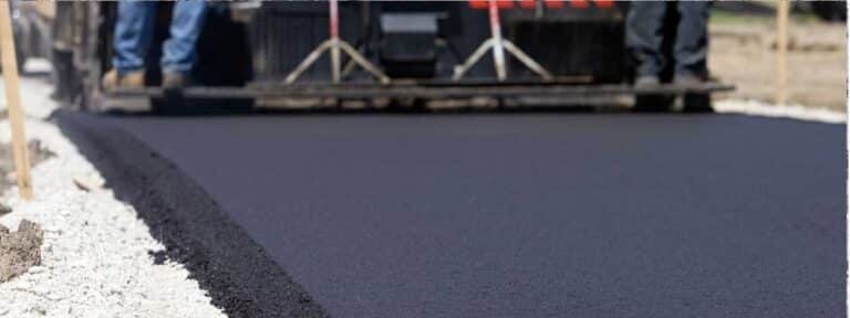 Close-up of fresh black asphalt being laid by paving machinery, with workers' legs in the background and gravel on the side—expertly handled by a paving contractor serving Mercer County NJ & Bucks County PA.