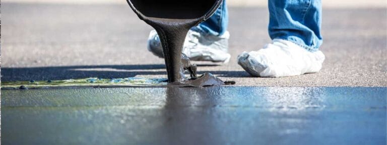 A person wearing jeans and white shoe covers pours black asphalt sealer onto a paved surface from a container, demonstrating the expertise of a paving contractor Hunterdon trusted by clients in Mercer County NJ & Bucks County PA.