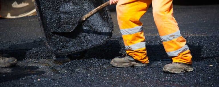 A construction worker in orange reflective pants and black boots spreads hot asphalt on a road with a shovel—exemplifying the skill of a paving contractor Hunterdon trusted across Mercer County NJ & Bucks County PA.