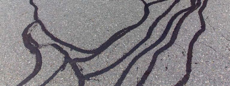 Dark liquid streaks are spread across a rough, gray asphalt surface, creating irregular, wavy lines and shapes—evidence of recent work by a paving contractor in Hunterdon, Mercer County NJ & Bucks County PA.